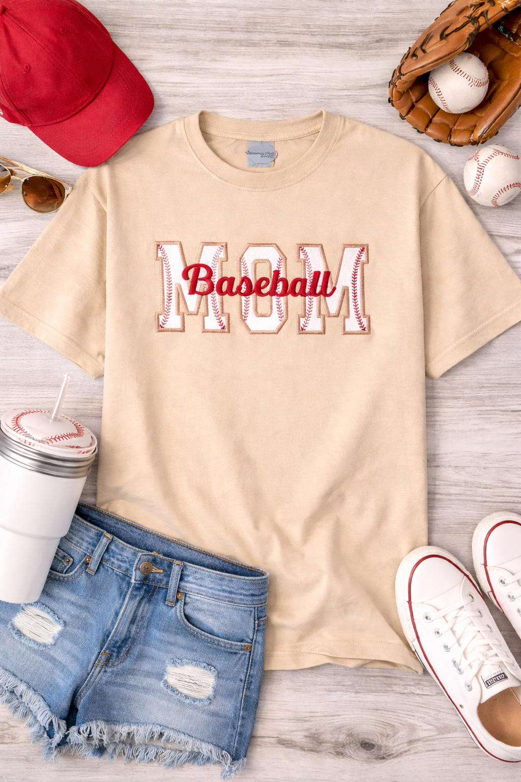 Beige t-shirt with 'Mom Baseball' text, red cap, sunglasses, shorts, sneakers, and baseballs on a wooden surface.
