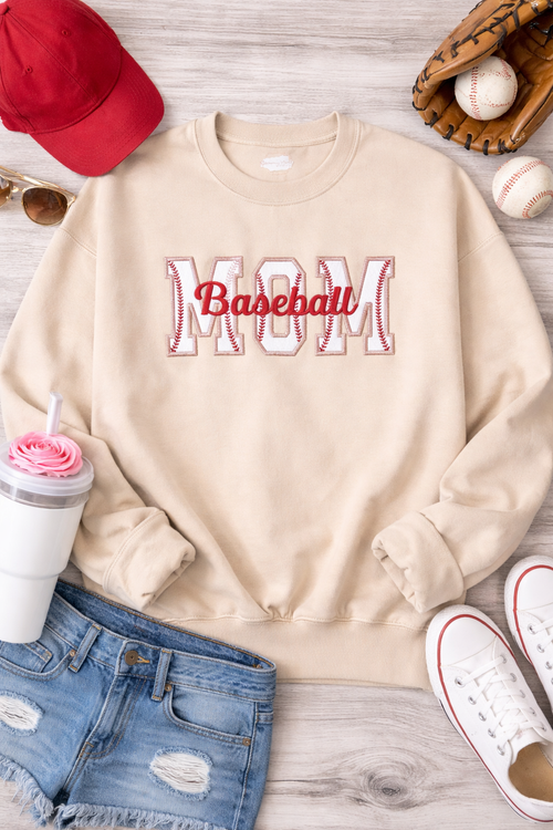 Beige sweatshirt with 'Mom Baseball' text, red cap, baseball glove, and sneakers on a wooden surface.