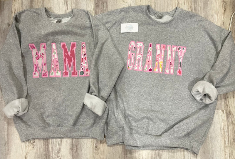 Two gray sweatshirts with 'MAMA' and 'GRANNY' text on a wooden surface