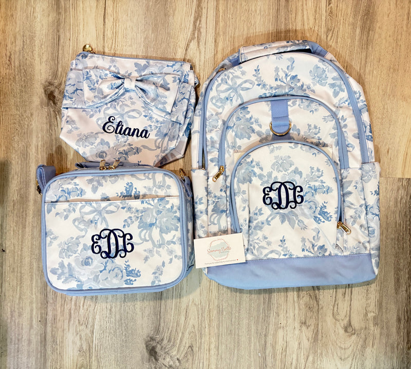 Floral-patterned backpack, lunch bag, and diaper bag set on a wooden surface with a embroidered monogram 