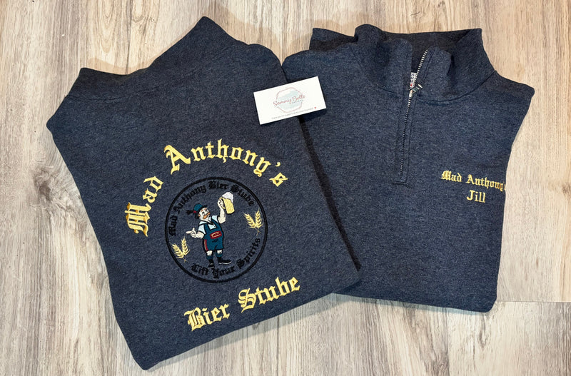 Two dark gray sweatshirts with gold text and logo on a wooden surface