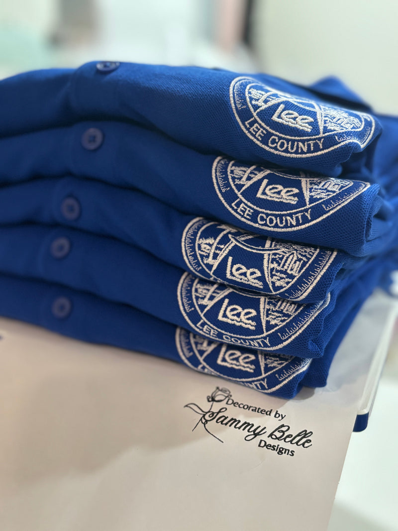 Stack of blue shirts with Lee County embroidered logo branding on a white surface.